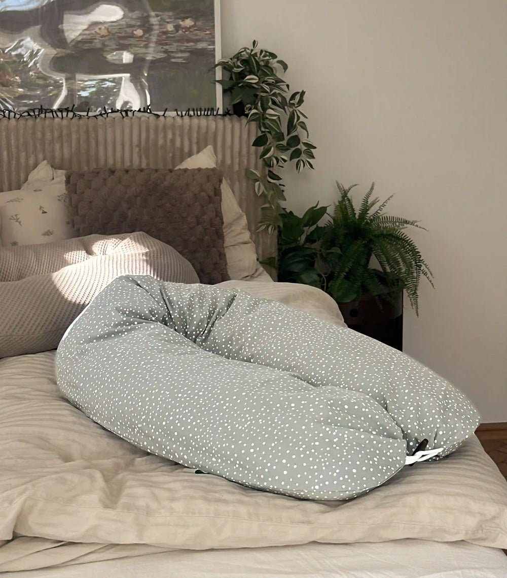 Ein Kreativkopf Still- und Seitenschläferkissen in einem glatten Bezug in grün liegt auf einem gemütlichen Bett. Der Holzknopf ist geschlossen. Im Hintergrund sieht man einige Pflanzen und ein weiteres Stillkissen