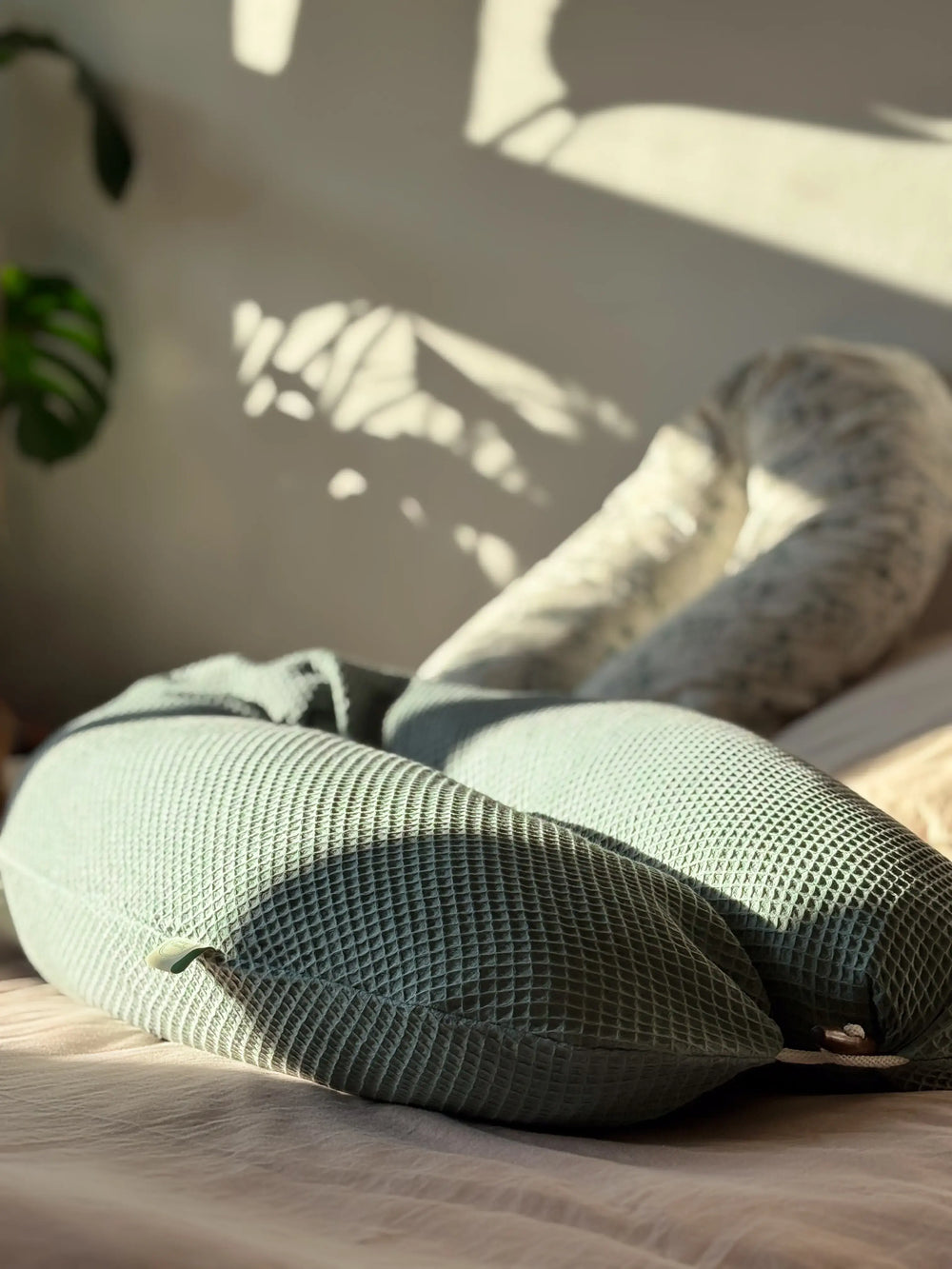 Ein Kreativkopf Still- und Seitenschläferkissen in einem Waffelbezug in grün liegt auf einem gemütlichen Bett. Der Holzknopf ist geschlossen. Im Hintergrund sieht man einige Pflanzen und ein weiteres Stillkissen
